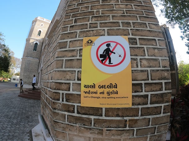 A large brick building with a sign on the wall showing a prohibition against spitting, featuring text in Gujarati and English. A man with a briefcase inside a circle with a line through it indicates the no-spitting rule. The background includes a tower with arched windows, trees, and people walking nearby.