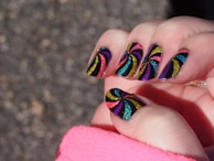 A close-up of a hand with nails painted in a vibrant, glittery spiral pattern. The design features colors like yellow, purple, blue, red, and green against a black background. The spiral patterns are bold and eye-catching, with each nail showcasing a similar but slightly varied design. A pink sleeve partially covers the wrist, suggesting the person is wearing a pink garment.