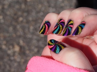 A close-up of a hand with nails painted in a vibrant, glittery spiral pattern. The design features colors like yellow, purple, blue, red, and green against a black background. The spiral patterns are bold and eye-catching, with each nail showcasing a similar but slightly varied design. A pink sleeve partially covers the wrist, suggesting the person is wearing a pink garment.