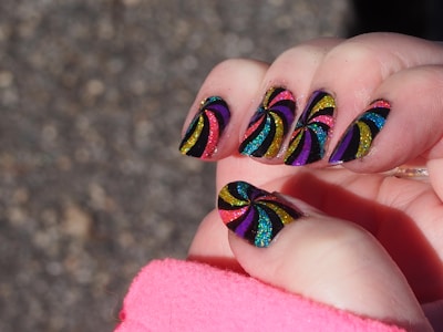 A close-up of a hand with nails painted in a vibrant, glittery spiral pattern. The design features colors like yellow, purple, blue, red, and green against a black background. The spiral patterns are bold and eye-catching, with each nail showcasing a similar but slightly varied design. A pink sleeve partially covers the wrist, suggesting the person is wearing a pink garment.