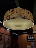 Large circular ceiling light with a lampshade covered in manga-style drawings. Wooden walls with signage and posters are visible, along with several smaller black light fixtures. The lighting creates a warm and cozy atmosphere.