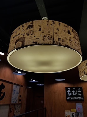 Large circular ceiling light with a lampshade covered in manga-style drawings. Wooden walls with signage and posters are visible, along with several smaller black light fixtures. The lighting creates a warm and cozy atmosphere.