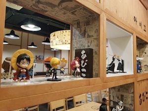 A collection of anime figurines is displayed on wooden shelves with comic-style wallpaper as a decorative background. Various action figures are posed, featuring vibrant colors and iconic character designs. The setting appears to be a themed cafe or restaurant, with additional seating and hanging lights in the background.