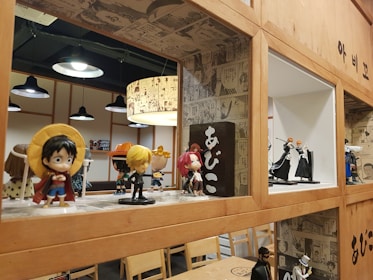 A collection of anime figurines is displayed on wooden shelves with comic-style wallpaper as a decorative background. Various action figures are posed, featuring vibrant colors and iconic character designs. The setting appears to be a themed cafe or restaurant, with additional seating and hanging lights in the background.