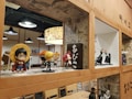 A collection of anime figurines is displayed on wooden shelves with comic-style wallpaper as a decorative background. Various action figures are posed, featuring vibrant colors and iconic character designs. The setting appears to be a themed cafe or restaurant, with additional seating and hanging lights in the background.