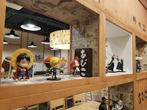 A collection of anime figurines is displayed on wooden shelves with comic-style wallpaper as a decorative background. Various action figures are posed, featuring vibrant colors and iconic character designs. The setting appears to be a themed cafe or restaurant, with additional seating and hanging lights in the background.
