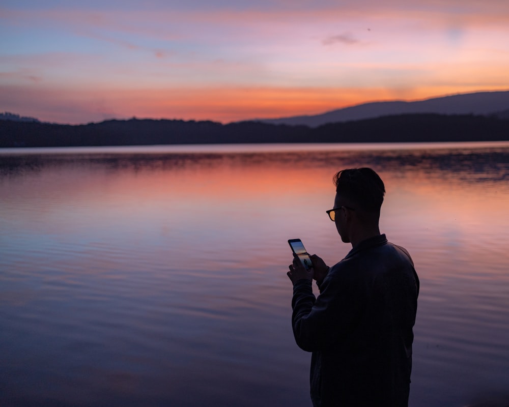 homme en veste noire tenant un smartphone près d’un plan d’eau au coucher du soleil