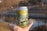 A hand holds a can with a detailed design featuring text that reads 'Sweet Dreams & Time Machines.' The illustration on the can depicts white lotus flowers against a serene backdrop of green foliage and sky. In the background, there's a blurred outdoor scene with a body of water and trees.
