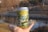 A hand holds a can with a detailed design featuring text that reads 'Sweet Dreams & Time Machines.' The illustration on the can depicts white lotus flowers against a serene backdrop of green foliage and sky. In the background, there's a blurred outdoor scene with a body of water and trees.