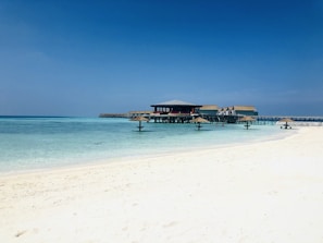 A serene beachfront scene with a clear blue sky overhead. The turquoise sea gently laps against the white sandy shore. A series of overwater bungalows on stilts stretch out into the ocean, connected by wooden walkways. There are a few small parasol-like structures on the water, providing a touch of shade.
