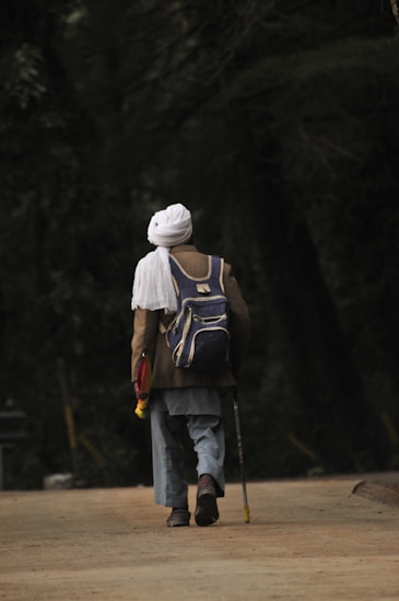 An elderly person is walking down a pathway surrounded by dense greenery. They are wearing a white turban, a brown coat, and carrying a backpack. The person is using a walking stick, indicating slow movement and support.