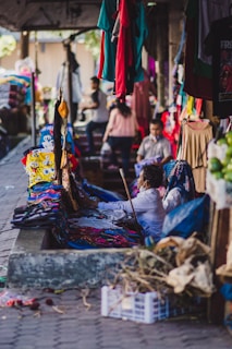 A bustling market scene with garments hanging and displayed on tables. People are interacting and shopping, creating a vibrant atmosphere. Various colors of clothing are noticeable, with some individuals focused on examining the merchandise.