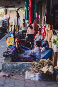 A bustling market scene with garments hanging and displayed on tables. People are interacting and shopping, creating a vibrant atmosphere. Various colors of clothing are noticeable, with some individuals focused on examining the merchandise.