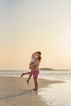 A couple is on a sandy beach at sunset, with the man lifting the woman while they embrace affectionately. The ocean is calm in the background, and the sky is tinged with soft hues from the setting sun.