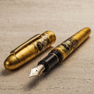 A luxurious fountain pen with intricate gold detailing featuring artistic engravings on the body. The pen is placed on a textured surface, and the cap is detached, displaying the polished nib.