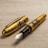 A luxurious fountain pen with intricate gold detailing featuring artistic engravings on the body. The pen is placed on a textured surface, and the cap is detached, displaying the polished nib.
