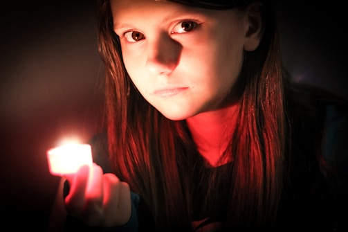 A person, possibly female, holds a glowing candle in a dimly lit setting. Their facial features are illuminated by the warm light of the candle, creating a contrast between the dark background and their face.