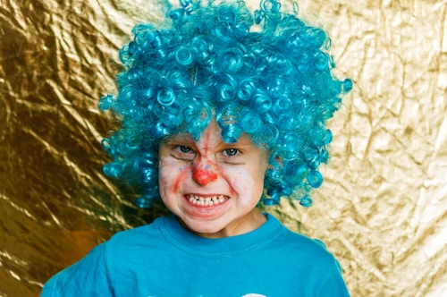 A child with a bright blue curly wig and clown makeup, featuring a red nose and exaggerated facial expressions. The background is a shiny gold texture, adding a playful and festive atmosphere.