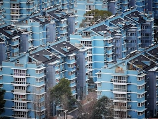 A densely packed residential area with multiple mid-rise apartments painted in various shades of blue. The buildings display modern architecture with repetitive geometric patterns, featuring several balconies and windows. Some trees and greenery are visible among the buildings, while the presence of air conditioning units is noticeable on the exterior walls.