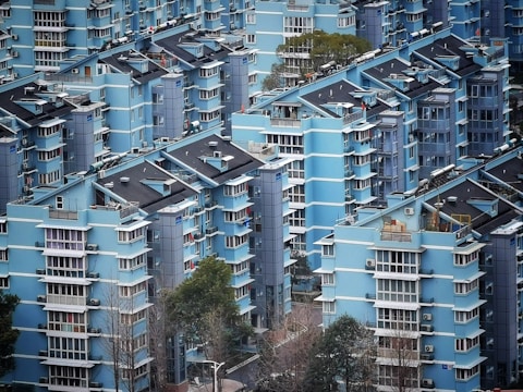 A densely packed residential area with multiple mid-rise apartments painted in various shades of blue. The buildings display modern architecture with repetitive geometric patterns, featuring several balconies and windows. Some trees and greenery are visible among the buildings, while the presence of air conditioning units is noticeable on the exterior walls.