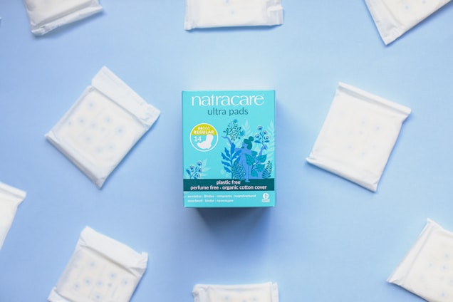 A package of Natracare ultra pads is centered on a light blue background, surrounded by individual sealed pads arranged in a circular manner. The packaging is a vibrant blue with floral illustrations and product information indicating features like plastic-free and organic cotton cover.