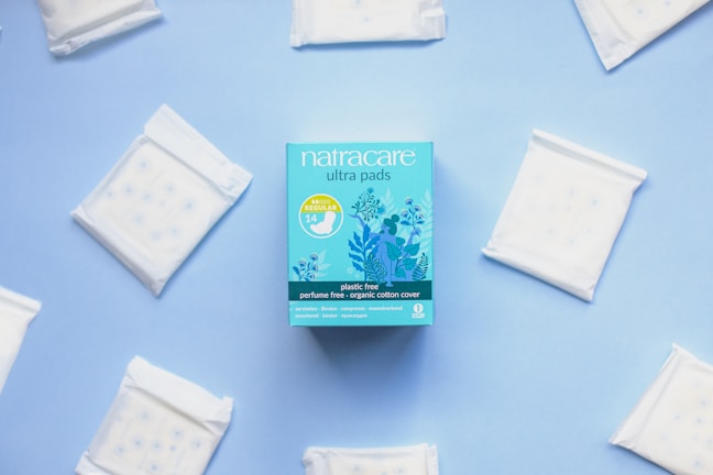 A package of Natracare ultra pads is centered on a light blue background, surrounded by individual sealed pads arranged in a circular manner. The packaging is a vibrant blue with floral illustrations and product information indicating features like plastic-free and organic cotton cover.