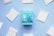 A package of Natracare ultra pads is centered on a light blue background, surrounded by individual sealed pads arranged in a circular manner. The packaging is a vibrant blue with floral illustrations and product information indicating features like plastic-free and organic cotton cover.