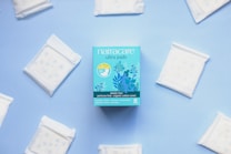 A package of Natracare ultra pads is centered on a light blue background, surrounded by individual sealed pads arranged in a circular manner. The packaging is a vibrant blue with floral illustrations and product information indicating features like plastic-free and organic cotton cover.