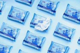 Multiple packets of Natracare intimate wipes arranged on a light blue background, with one open packet revealing a wipe. The packages are predominantly blue and feature floral designs in darker blue and yellow accents.