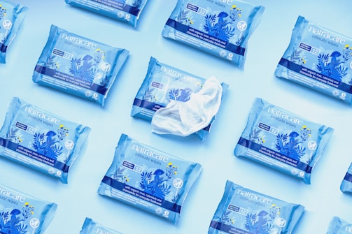 Multiple packets of Natracare intimate wipes arranged on a light blue background, with one open packet revealing a wipe. The packages are predominantly blue and feature floral designs in darker blue and yellow accents.