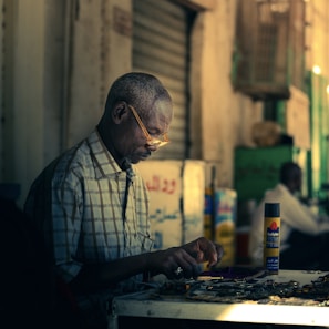 An elderly man, wearing glasses, sits at a workbench in a dimly lit environment. He appears focused as he works with small objects, possibly watches or other items, spread on the table in front of him. The background is blurred, highlighting the subject's concentration and dedication to his task. Light softly illuminates the area, creating a calm and focused atmosphere.