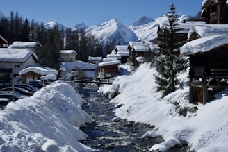 A picturesque snowy village nestled in a mountainous region. Wooden chalets line the snowy banks of a flowing river, surrounded by tall pine trees. In the background, snow-capped mountains are visible under a clear blue sky.