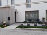 A modern, minimalist house exterior with a combination of wooden panels and white walls. The front features a small patio area with two black chairs and a small table between them. There are three large rectangular planters with green ornamental grass, and a flower pot with chrysanthemums and a pumpkin decorates the sidewalk. The entrance has a modern design with a black mailbox numbered 11.