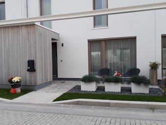 A modern, minimalist house exterior with a combination of wooden panels and white walls. The front features a small patio area with two black chairs and a small table between them. There are three large rectangular planters with green ornamental grass, and a flower pot with chrysanthemums and a pumpkin decorates the sidewalk. The entrance has a modern design with a black mailbox numbered 11.