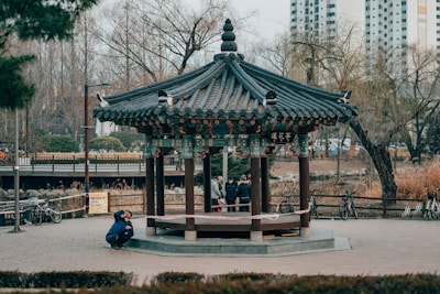 man in black jacket sitting on brown wooden bench near green and brown temple during daytime