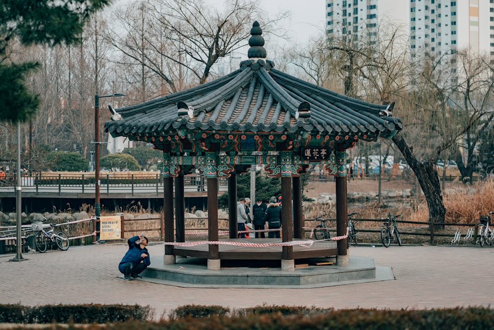 Hombre en chaqueta negra sentado en banco de madera marrón cerca del templo verde y marrón durante el día