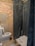 A rustic bathroom scene with exposed brick and concrete walls. A white toilet is positioned on the left, with an open lid. To the right is a shower area covered with a dark, textured curtain. Various toiletries, including a green bottle and a tube, are placed near the shower.