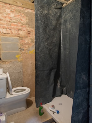 A rustic bathroom scene with exposed brick and concrete walls. A white toilet is positioned on the left, with an open lid. To the right is a shower area covered with a dark, textured curtain. Various toiletries, including a green bottle and a tube, are placed near the shower.