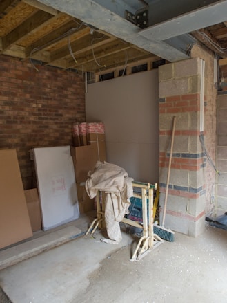 A partially constructed room with exposed brick and wooden beams. Building materials like boards and cardboard boxes are stacked against the walls. Scaffolding supports are placed in the middle, draped with a cloth. The floor is concrete, and tools or equipment can be seen around.