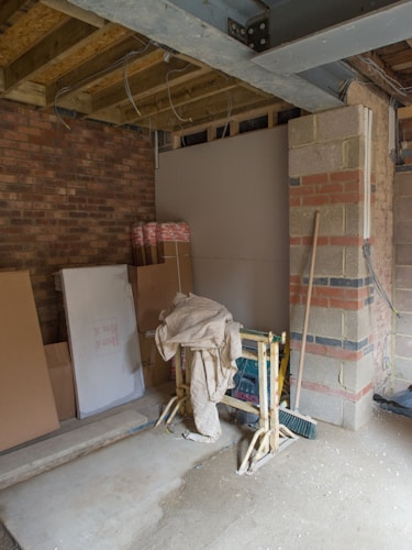 A partially constructed room with exposed brick and wooden beams. Building materials like boards and cardboard boxes are stacked against the walls. Scaffolding supports are placed in the middle, draped with a cloth. The floor is concrete, and tools or equipment can be seen around.