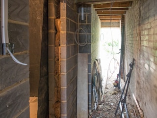 A narrow construction space with exposed brick and block walls, scattered construction materials, and some wiring hanging from the ceiling. The space appears unfinished, with debris on the ground and light streaming in from an opening at the end.