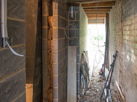A narrow construction space with exposed brick and block walls, scattered construction materials, and some wiring hanging from the ceiling. The space appears unfinished, with debris on the ground and light streaming in from an opening at the end.