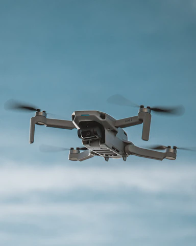 A quadcopter drone is captured in mid-air against a clear blue sky. The drone's propellers are spinning, visible as slightly blurred from motion. It is composed of a sleek grey body with visible camera gimbal underneath, designed for aerial photography.