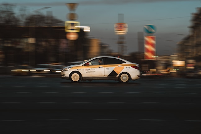 A taxi car in motion with a blurred city background, capturing a sense of speed and movement. The taxi is white with yellow and black markings and is driving along a street with various urban elements such as road signs visible.