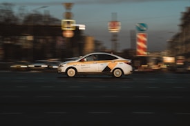 A taxi car in motion with a blurred city background, capturing a sense of speed and movement. The taxi is white with yellow and black markings and is driving along a street with various urban elements such as road signs visible.