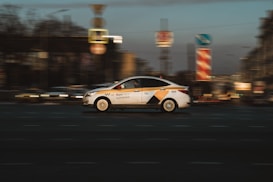 A taxi car in motion with a blurred city background, capturing a sense of speed and movement. The taxi is white with yellow and black markings and is driving along a street with various urban elements such as road signs visible.