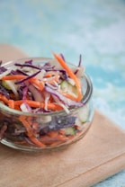A glass jar filled with a colorful salad containing shredded carrots, purple cabbage, and slices of cucumber. The background is an indistinct blend of blue and green tones, and the jar is resting on a light wooden surface.