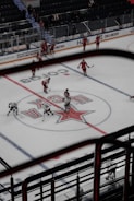 A hockey game is taking place in an indoor arena. Players in red and white uniforms are scattered across the ice, some holding hockey sticks. The central logo on the ice indicates the home team. The arena has stands with a few spectators visible. There are boards with advertisements around the perimeter of the rink.