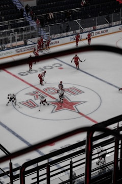 A hockey game is taking place in an indoor arena. Players in red and white uniforms are scattered across the ice, some holding hockey sticks. The central logo on the ice indicates the home team. The arena has stands with a few spectators visible. There are boards with advertisements around the perimeter of the rink.