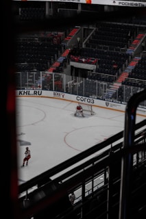 A hockey game taking place in an indoor arena with visible advertisements around the rink. The ice surface shows a goalie positioned in front of the net, and another player is near the boards wearing a red and white uniform. The arena seating is mostly empty, and there's a noticeable architectural element with steps leading up the stands.